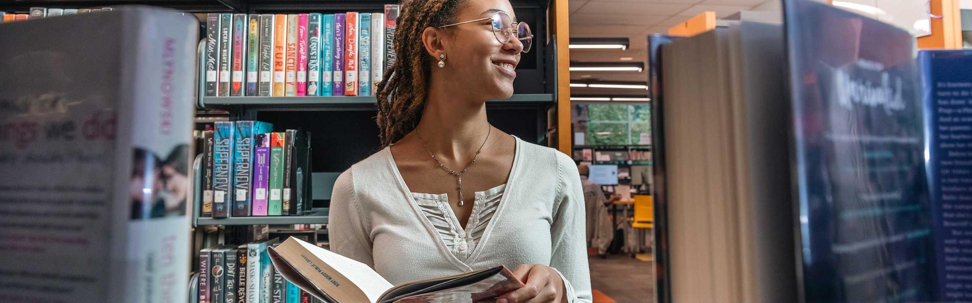 A young woman stands in from of library shelves, holding a book open.
